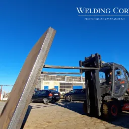 Pomarańczowy wózek widłowy Linde H45 transportuje długi, metalowy profil o przekroju prostokątnym na tle błękitnego nieba i budynku przemysłowego z rzędami okien.