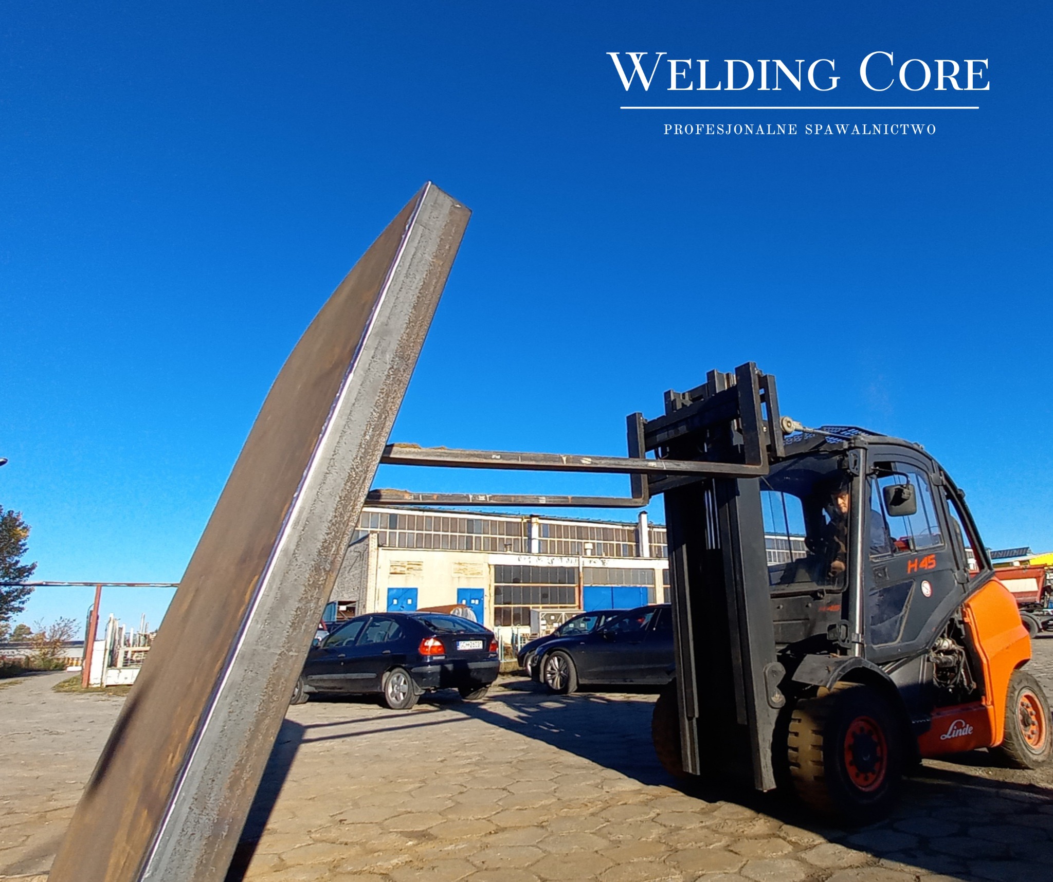 Pomarańczowy wózek widłowy Linde H45 transportuje długi, metalowy profil o przekroju prostokątnym na tle błękitnego nieba i budynku przemysłowego z rzędami okien.