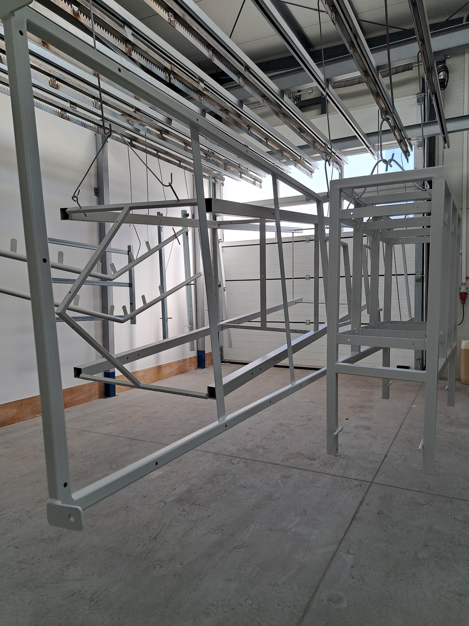 Metalowe ramy konstrukcyjne, pomalowane na szaro, zawieszone na hakach w hali produkcyjnej, widoczne szyny transportowe pod sufitem i brama w tle.