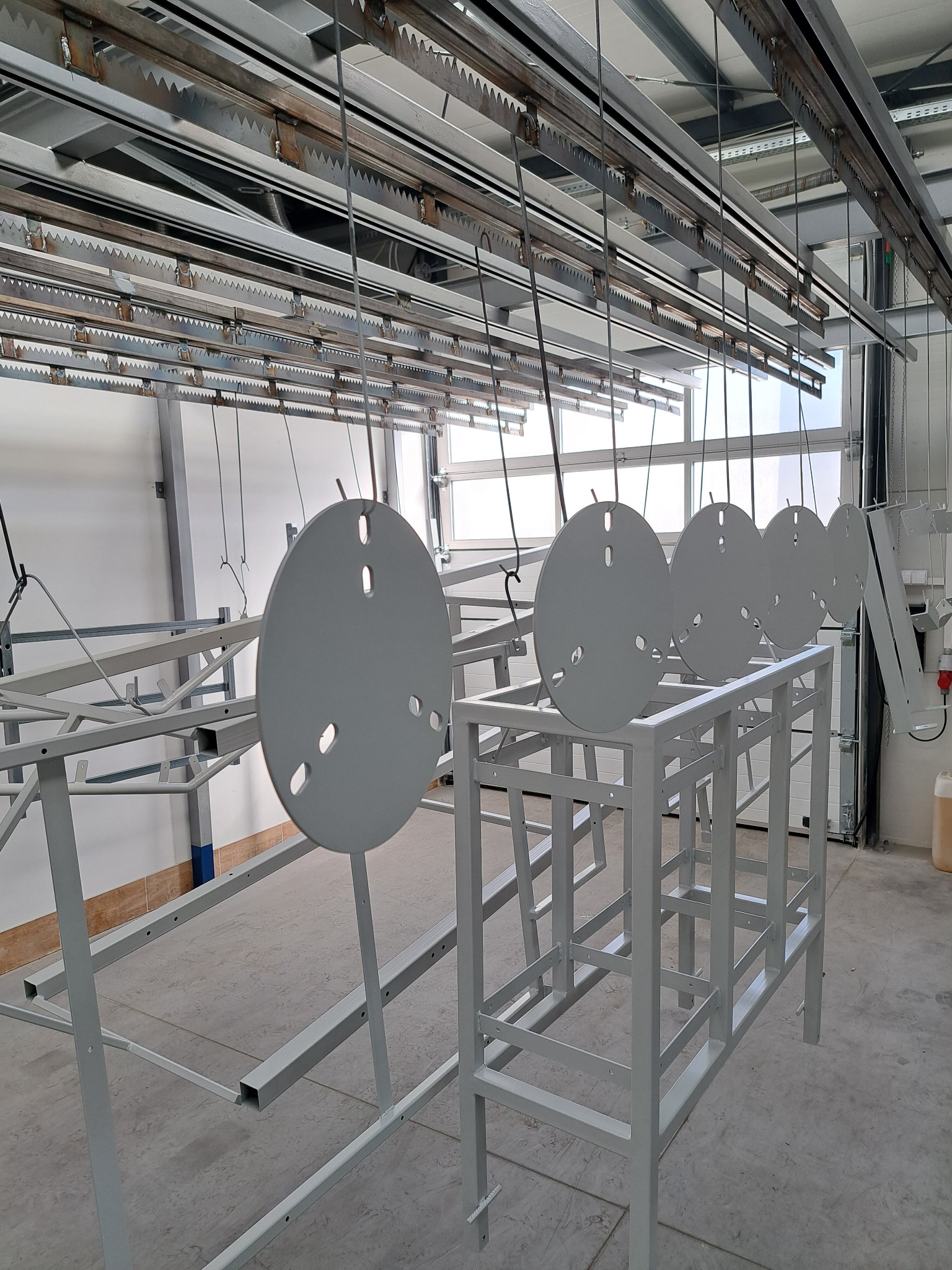 Szare, okrągłe elementy z otworami, zawieszone na hakach w hali produkcyjnej, prawdopodobnie po procesie lakierowania.