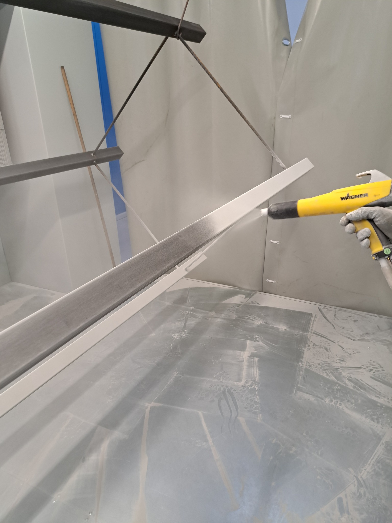 Proces malowania proszkowego elementu metalowego w kabinie lakierniczej, z widocznym pistoletem Wagner i efektem rozpylania farby.