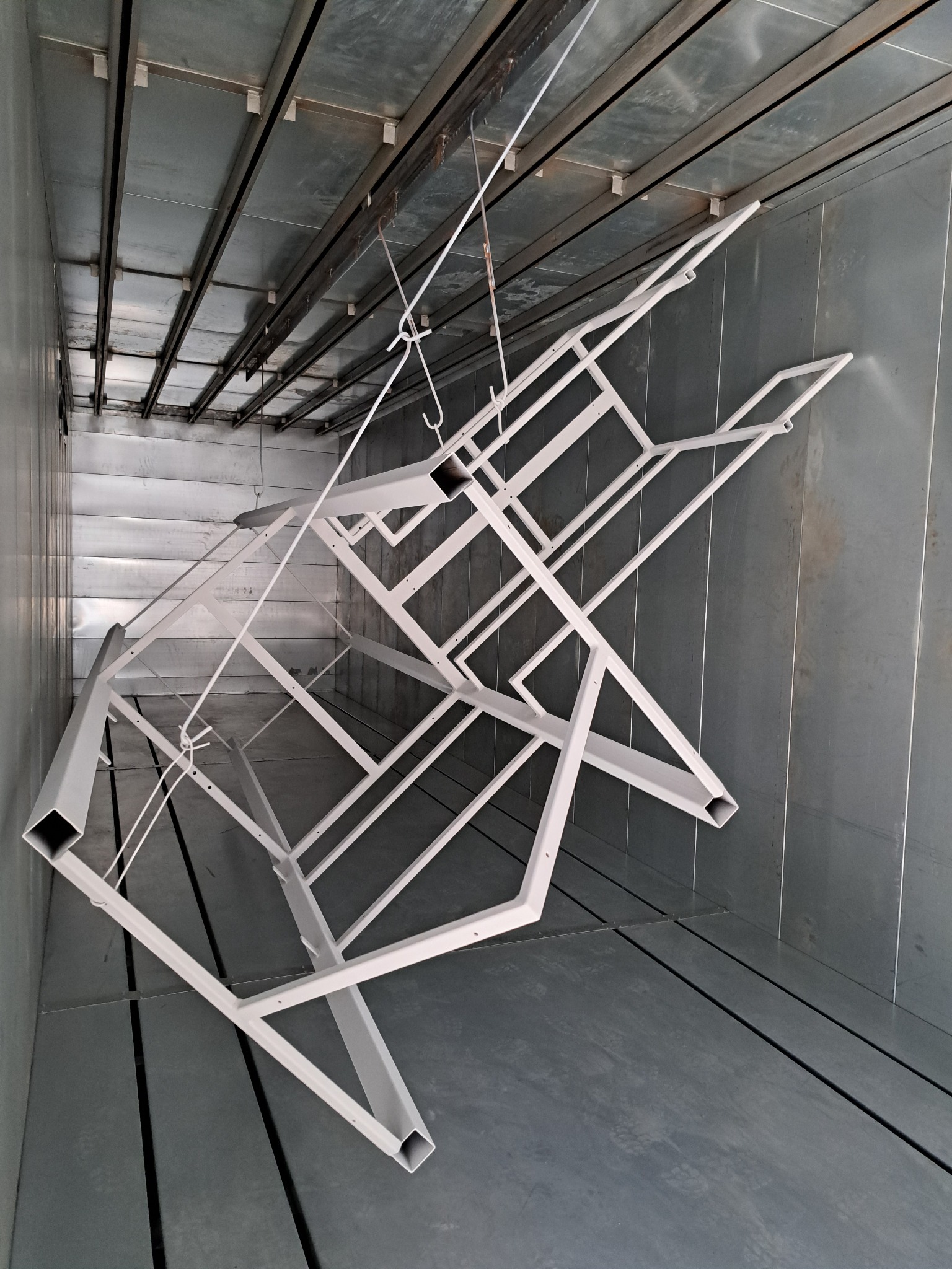 Metalowa konstrukcja ramy, zawieszona na hakach w komorze, świeżo po nałożeniu jasnoszarej powłoki.