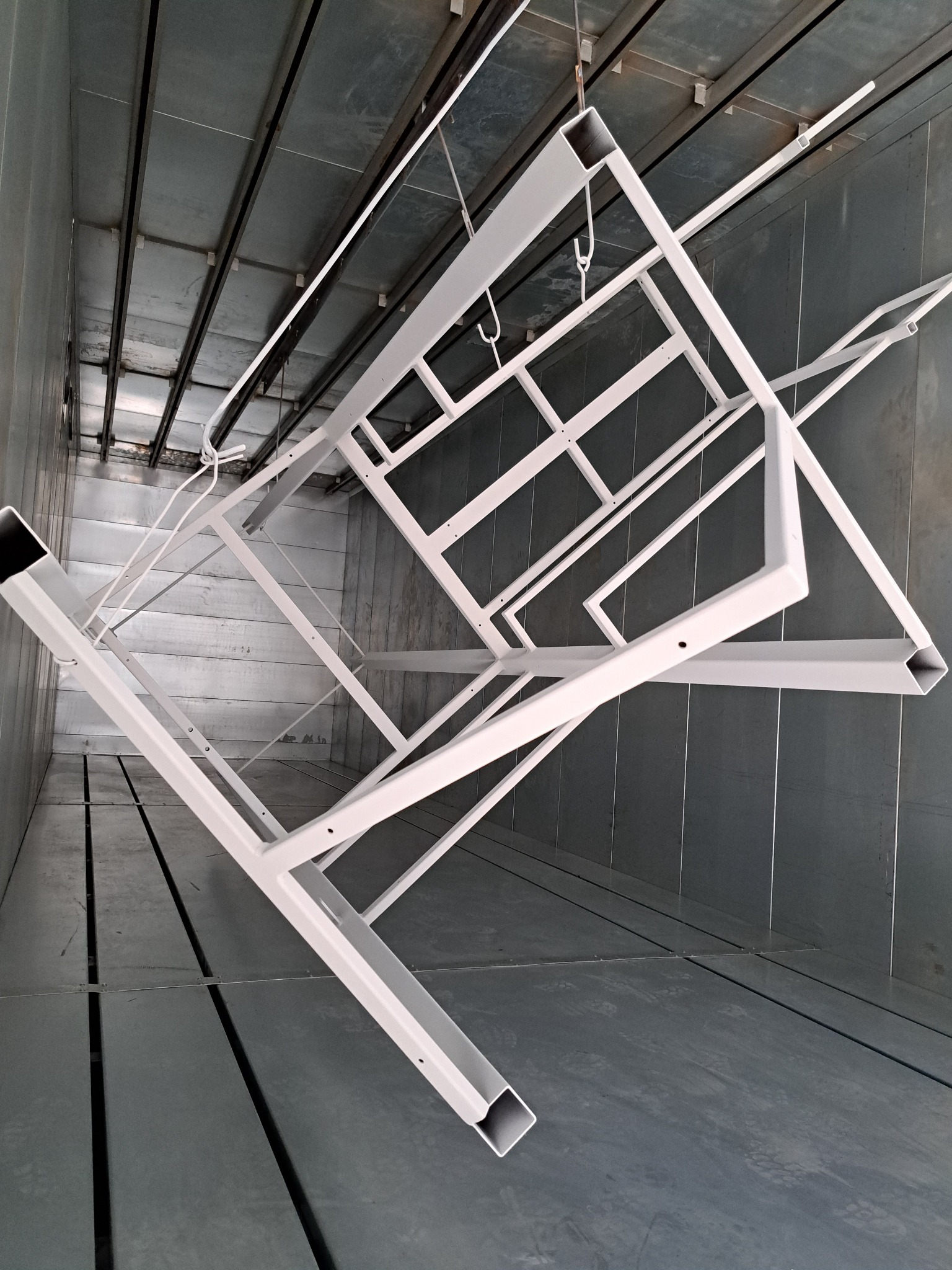 Metalowa konstrukcja wisząca na hakach wewnątrz lakierni proszkowej, świeżo po nałożeniu białej powłoki.