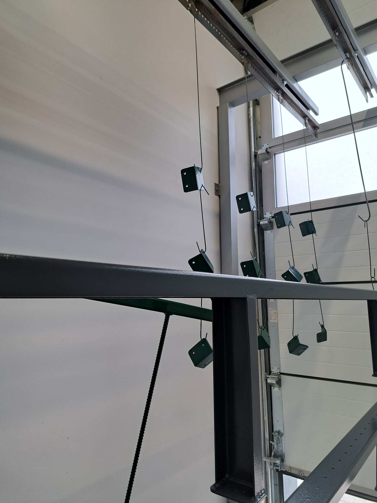 Elementy metalowe zawieszone na lince, przygotowane do malowania proszkowego, widoczne szyny transportowe i brama garażowa w tle.