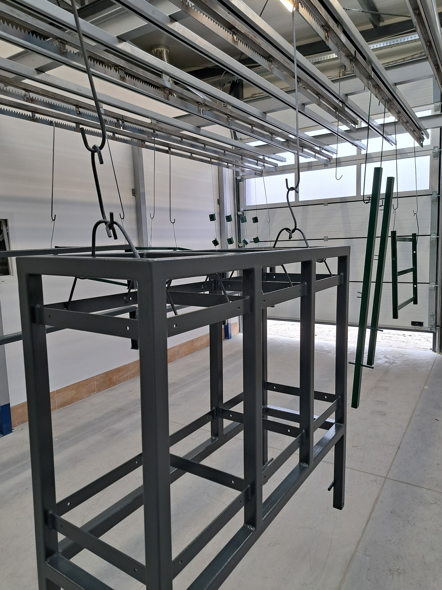 Metalowa konstrukcja stołu zawieszona na hakach w hali, przygotowana do dalszej obróbki lub po procesie malowania, widoczne elementy transportu surowców pod sufitem.