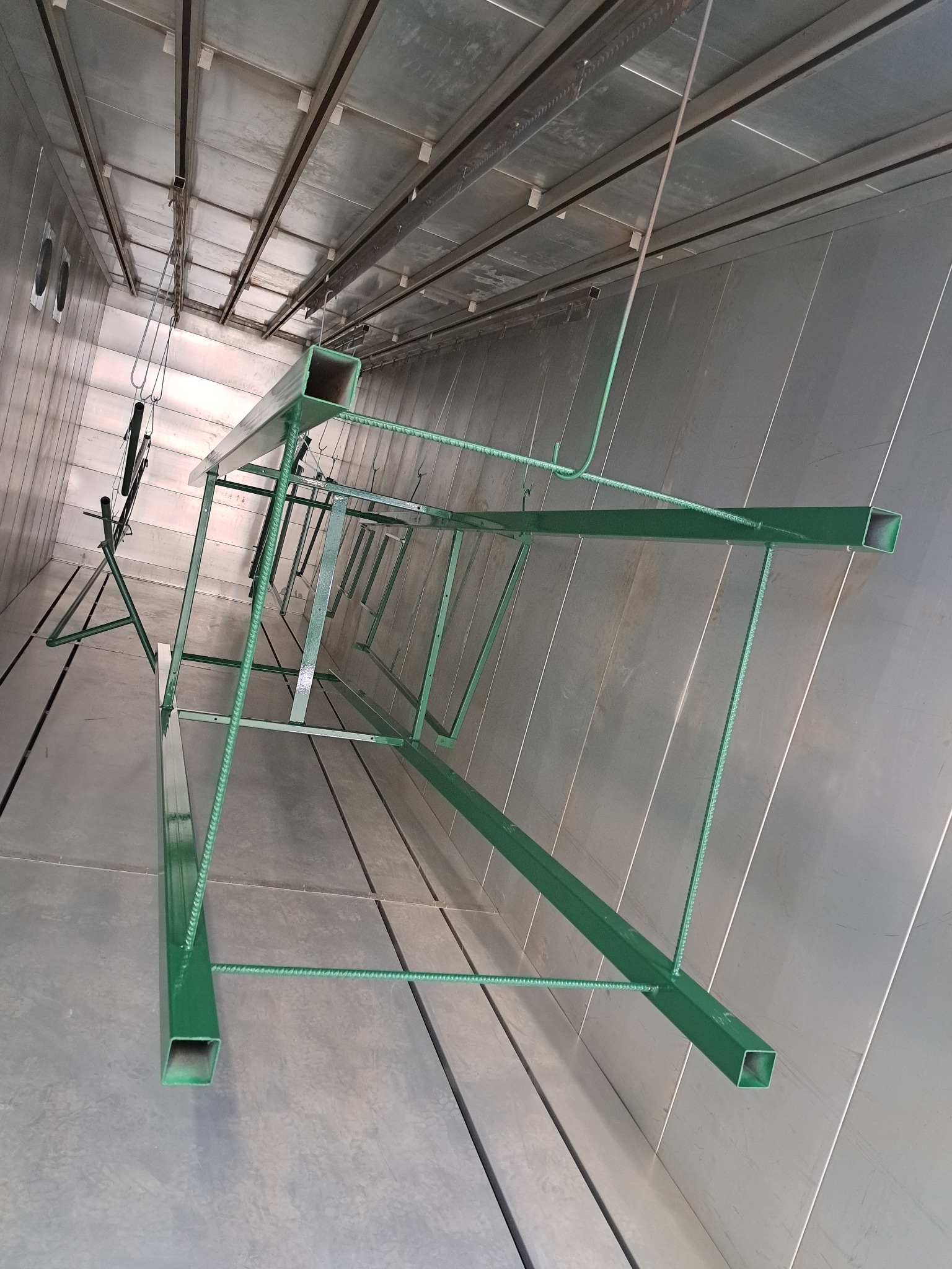 Metalowa konstrukcja pomalowana na zielono, wisząca wewnątrz przemysłowej kabiny lakierniczej, gotowa lub po procesie malowania.