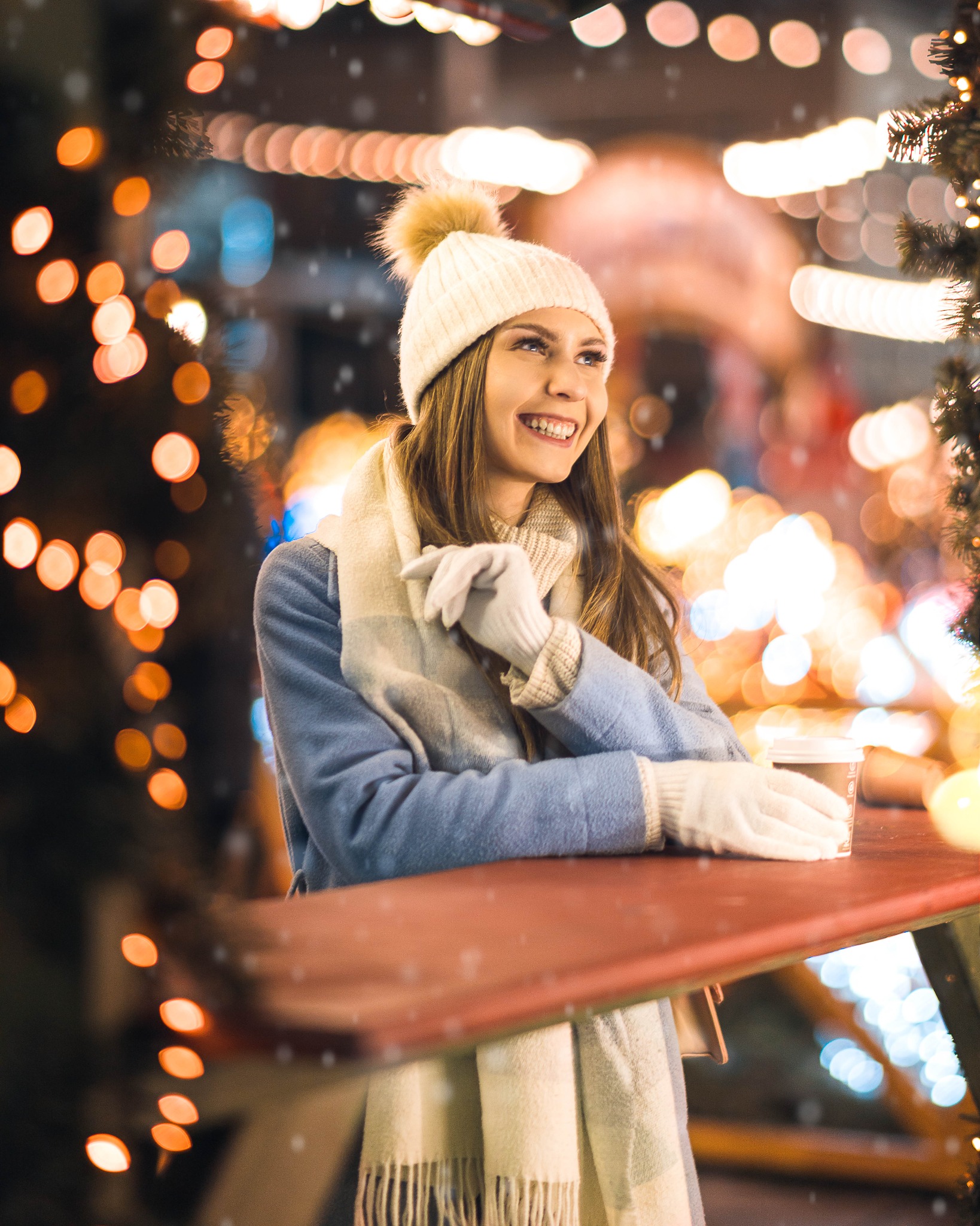 Uśmiechnięta młoda kobieta w zimowej czapce i szaliku, oparta o czerwony blat, z kubkiem kawy na tle świątecznych światełek.