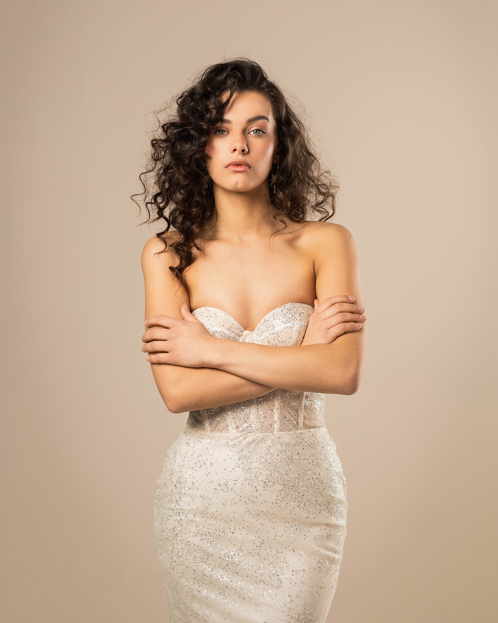 Portret kobiety z ciemnymi kręconymi włosami, ubranej w białą, błyszczącą suknię bez ramiączek, z założonymi rękami na piersiach, na jednolitym beżowym tle.
