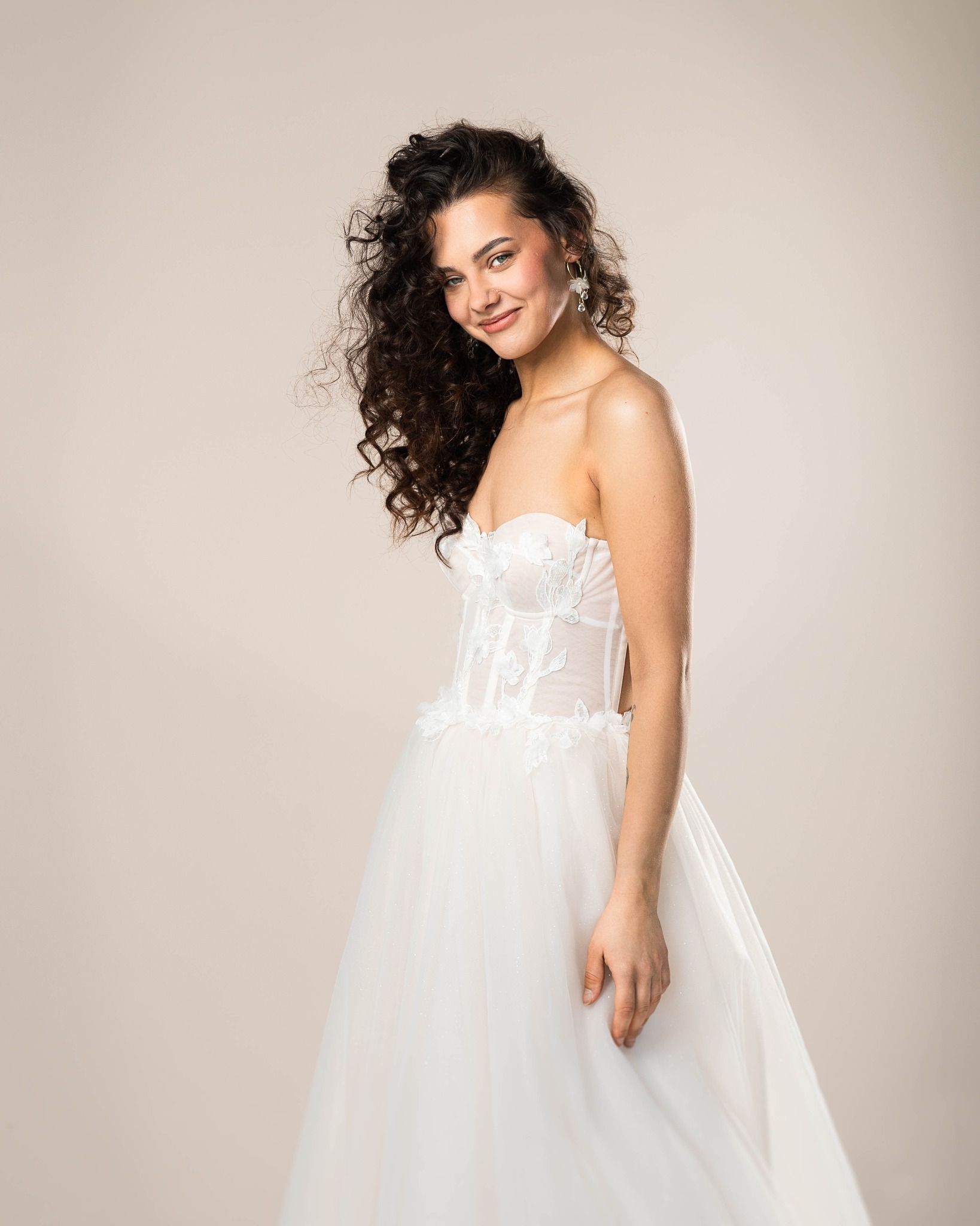 Uśmiechnięta panna młoda w białej sukni ślubnej z tiulowym dołem i kwiatowymi aplikacjami na gorsecie, pozująca na jasnym tle.