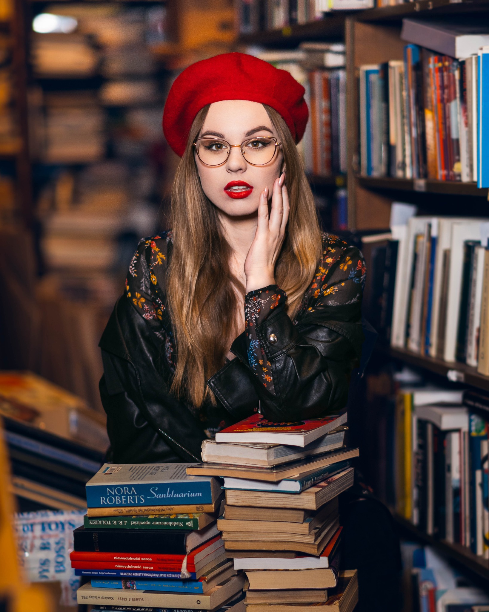 Portret kobiety w czerwonym berecie i okularach, opierającej się o stos książek w bibliotece, z ręką przy twarzy i mocnym makijażem.