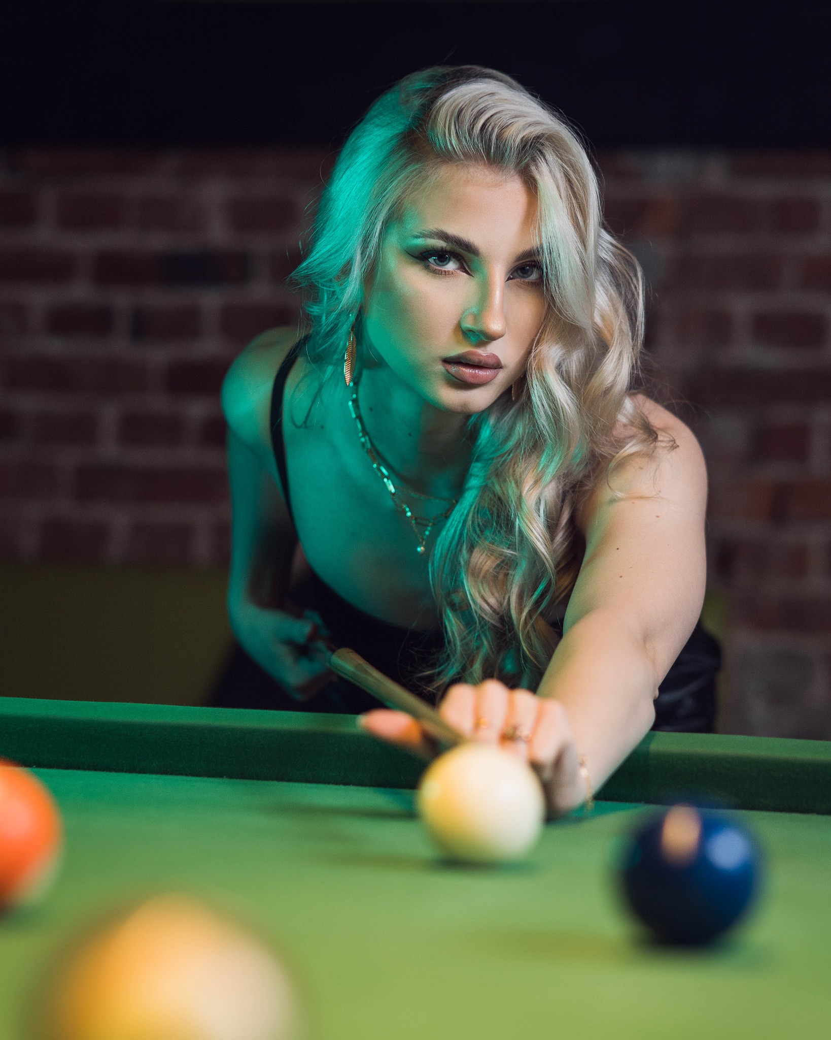 Kobieta z blond włosami, w biżuterii, celuje kijem bilardowym w białą bilę na zielonym stole bilardowym, ceglana ściana w tle.