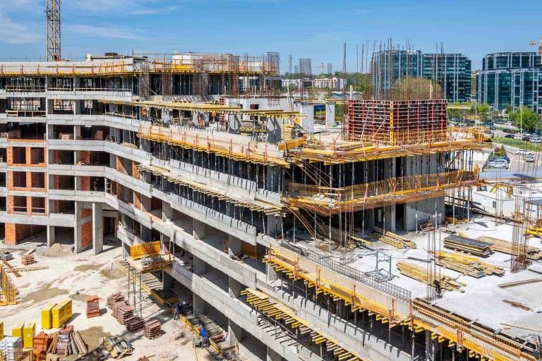 Wielopiętrowy budynek w stanie surowym, widok z góry na plac budowy z widocznymi elementami szalunków, zbrojenia i konstrukcji żelbetowej, w tle zabudowa miejska i błękitne niebo.