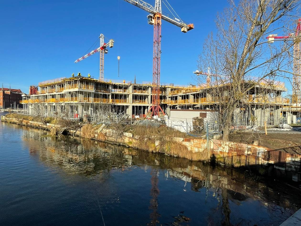 Wznoszenie wielopiętrowego budynku w stanie surowym, widok od strony lustra wody, z widocznymi żurawiami budowlanymi i pracownikami na rusztowaniach.