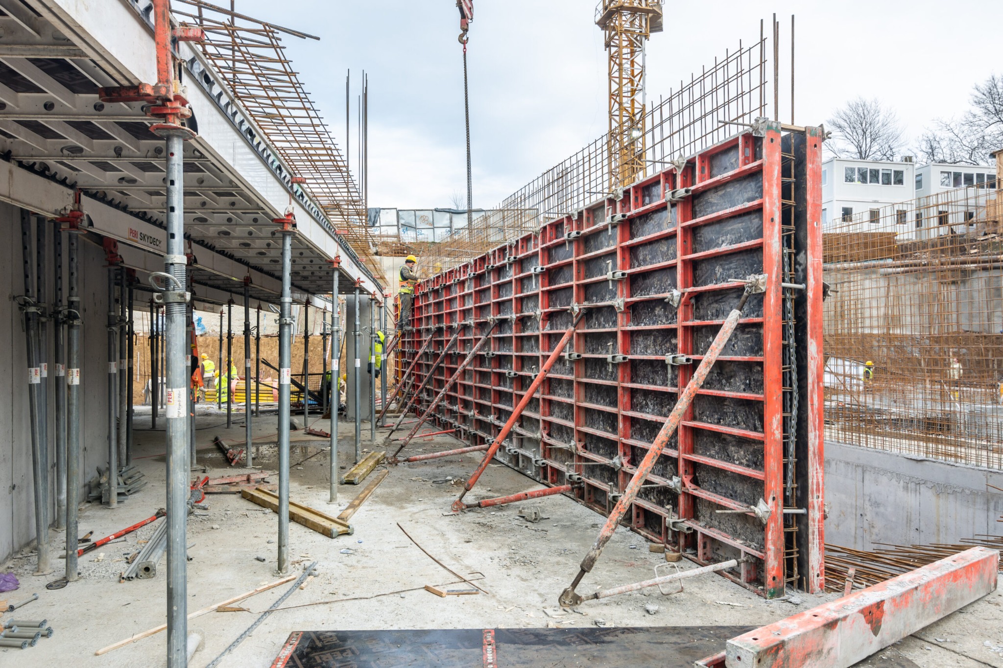 Szare betonowe fundamenty w trakcie budowy, z widocznymi deskowaniami w kolorze czerwonym i stalowymi zbrojeniami, wsparte na metalowych podporach. W tle pracownicy w żółtych kaskach i dźwig...