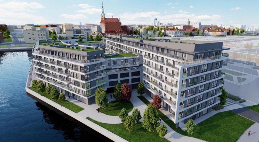 Wizualizacja kompleksu budynków żelbetowych z zielonymi dachami, położonych nad rzeką w tle miejskiego krajobrazu z kościołem.