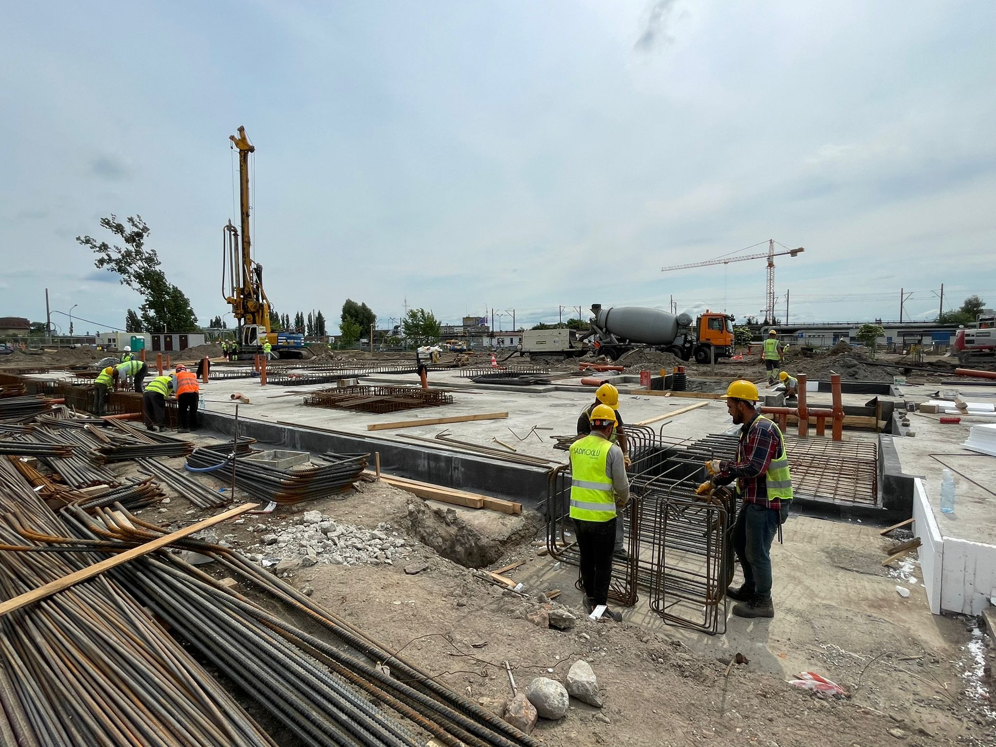 Szeroki plan budowy fundamentów z widoczną zbrojoną konstrukcją, pracownikami w żółtych kamizelkach i betoniarką w tle, na placu budowy widoczna wiertnica.