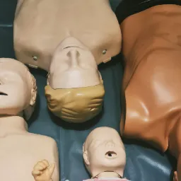 Manekiny do nauki pierwszej pomocy, w tym model niemowlęcia w różowo-białej koszulce, leżące na niebieskim podłożu, widoczne elementy urządzenia do wentylacji.