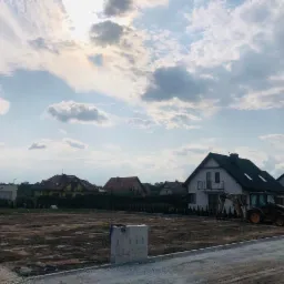 Przygotowany teren pod budowę osiedla domów jednorodzinnych, widoczny wyrównany grunt i pracująca koparko-ładowarka na tle zabudowy mieszkalnej pod pochmurnym niebem.