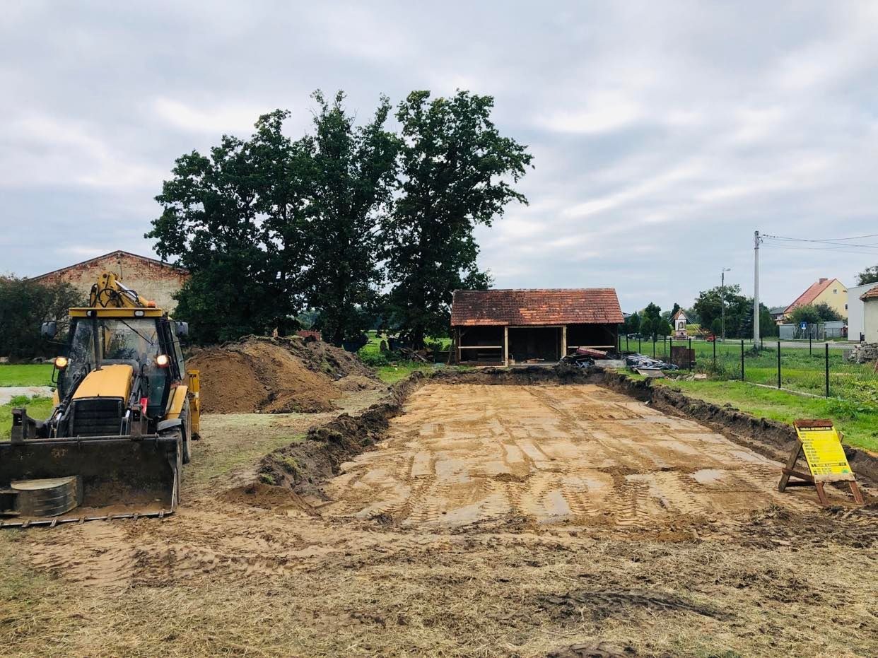 Żółta koparko-ładowarka na placu budowy, przygotowanie terenu pod budowę, widoczne ślady opon na piasku, w tle drewniana konstrukcja i drzewa.