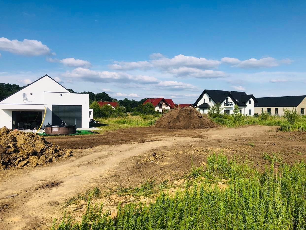 Przygotowany teren pod budowę z usypaną pryzmą ziemi, widoczne nowe domy w tle i nowa instalacja zewnętrznego jacuzzi przy garażu.