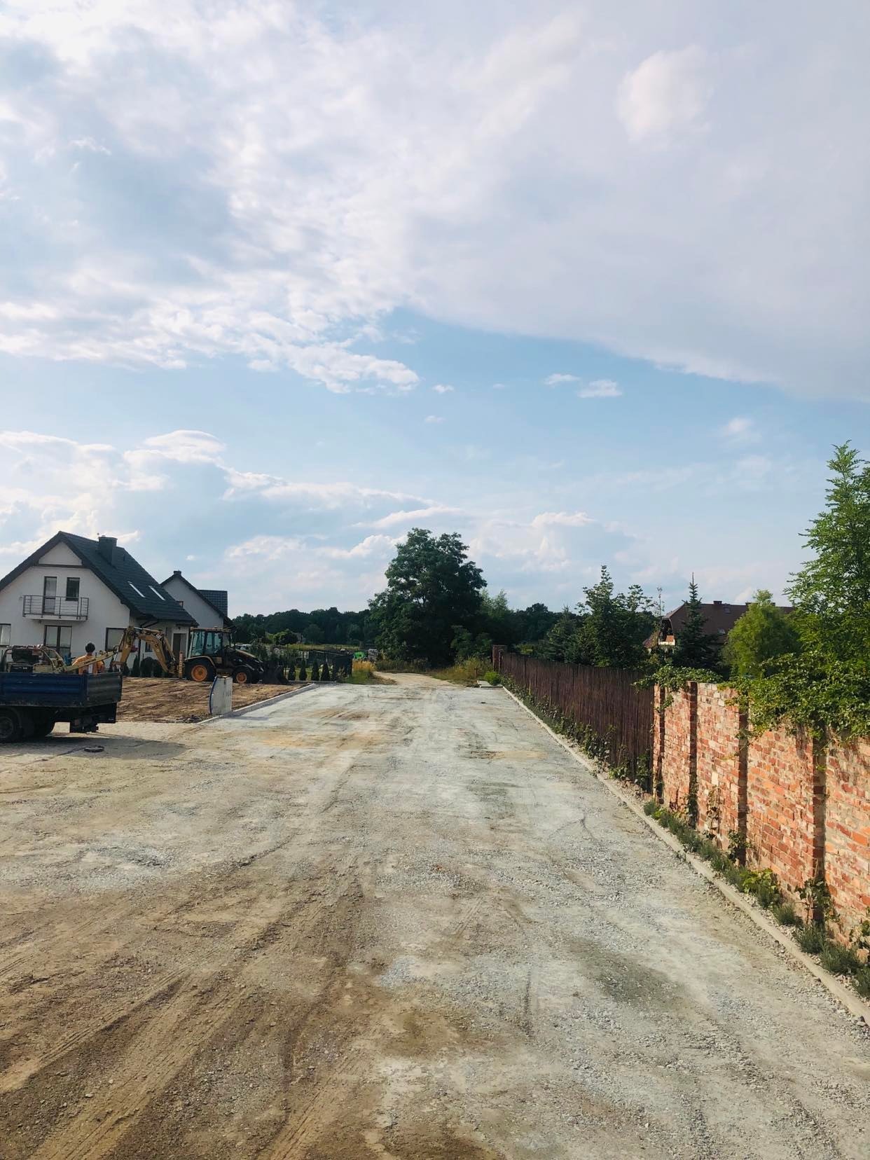 Przygotowany teren pod budowę drogi dojazdowej, widoczny sprzęt budowlany i częściowo ułożona podbudowa z kruszywa, obok ceglany mur i drewniane ogrodzenie.