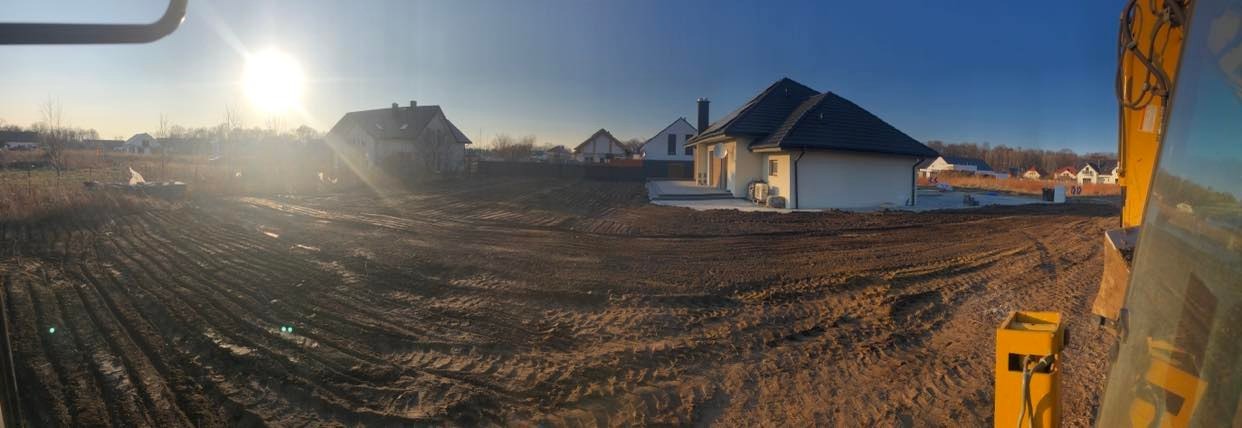 Przygotowany teren pod budowę domu w Kątach Wrocławskich, widok z kabiny koparki na wyrównaną działkę z widocznymi śladami opon i fundamentami pod budynek.