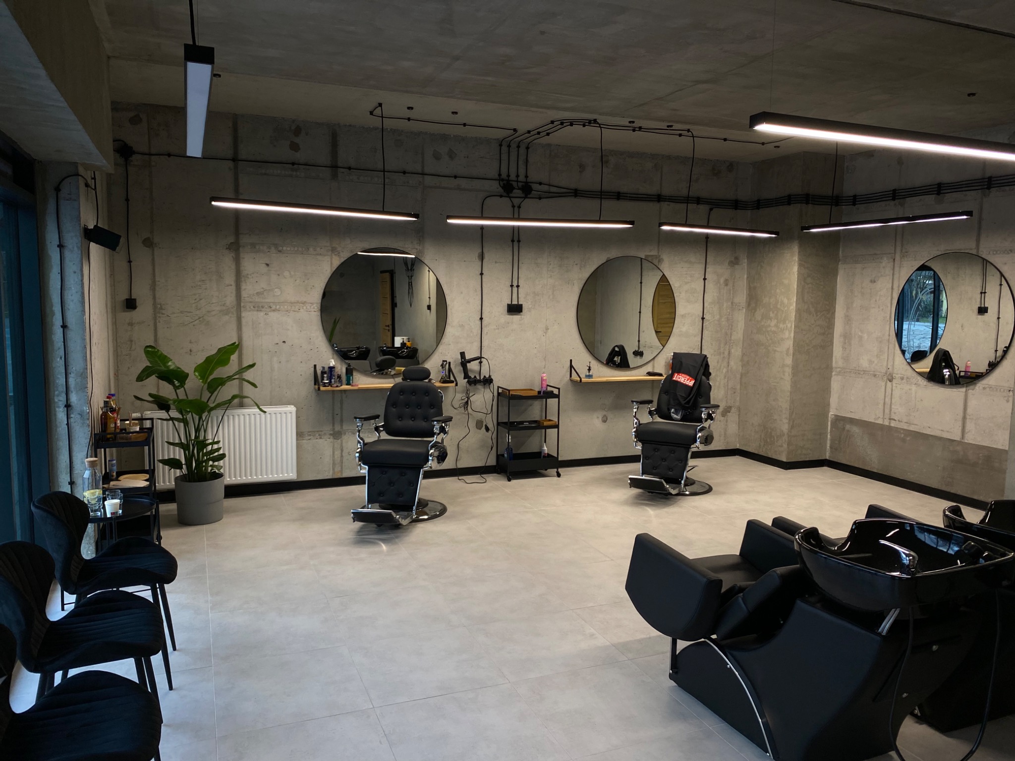 Nowoczesne wnętrze salonu z trzema stanowiskami fryzjerskimi, okrągłymi lustrami, betonowymi ścianami i minimalistycznym oświetleniem liniowym, uzupełnione rośliną doniczkową i czarnymi fotelami...