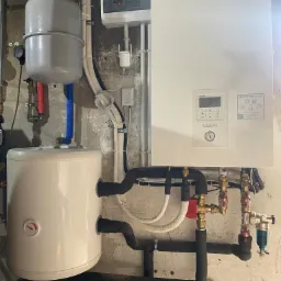 Biała pompa ciepła Panasonic Aquarea z podłączonym zbiornikiem i orurowaniem na tle surowej ściany z betonu.