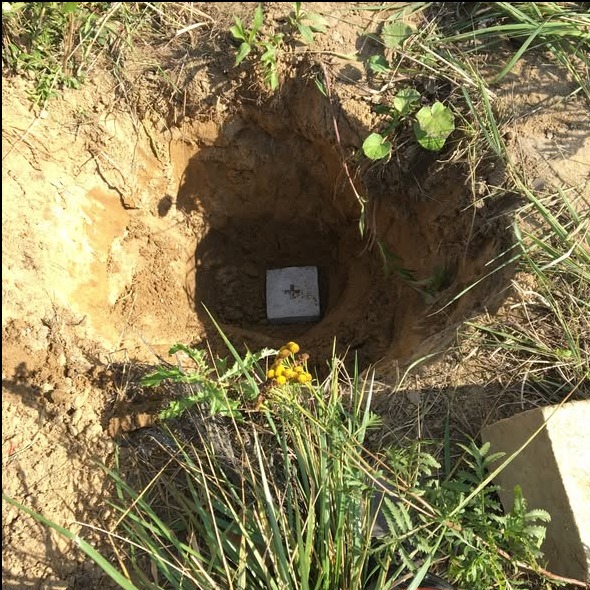 Betonowy słupek geodezyjny z krzyżem w wykopie, otoczony ziemią i zielenią. Widok z góry, naturalne światło.