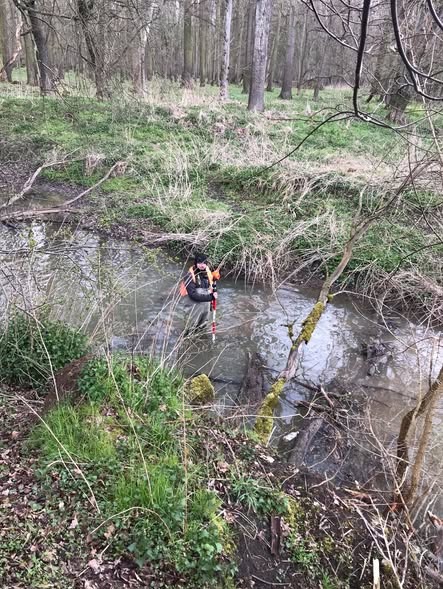 Osoba w kombinezonie wchodzi do płytkiego strumienia w lesie, trzymając tyczkę geodezyjną, prawdopodobnie wykonując pomiary w terenie zalesionym.