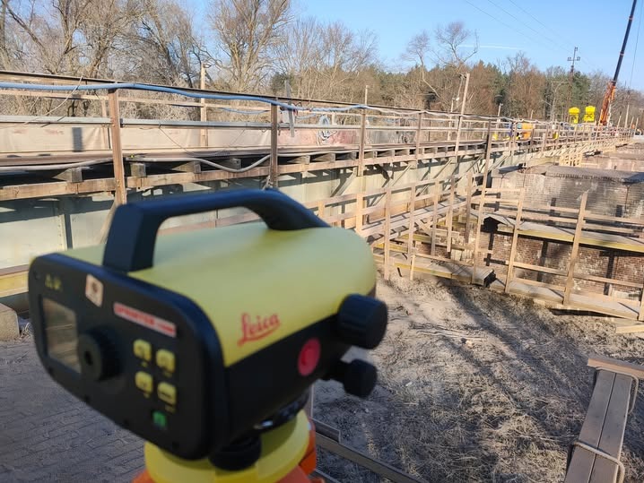 Żółty niwelator Leica na statywie, w tle konstrukcja mostu w trakcie budowy z drewnianym szalunkiem i piaskiem. Widoczny dźwig budowlany w oddali.