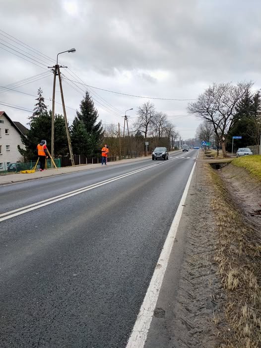 Praca na drodze: dwóch pracowników w pomarańczowych kamizelkach, używa sprzętu pomiarowego na poboczu ruchliwej ulicy w pochmurny dzień. Widoczne drzewa i budynki w tle.