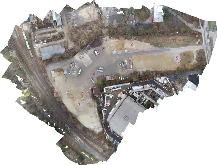 Szeroki, ukośny widok z drona na teren budowy z zaznaczonymi obszarami roboczymi, widoczne pojazdy i budynki w tle, teren częściowo przygotowany pod inwestycję.