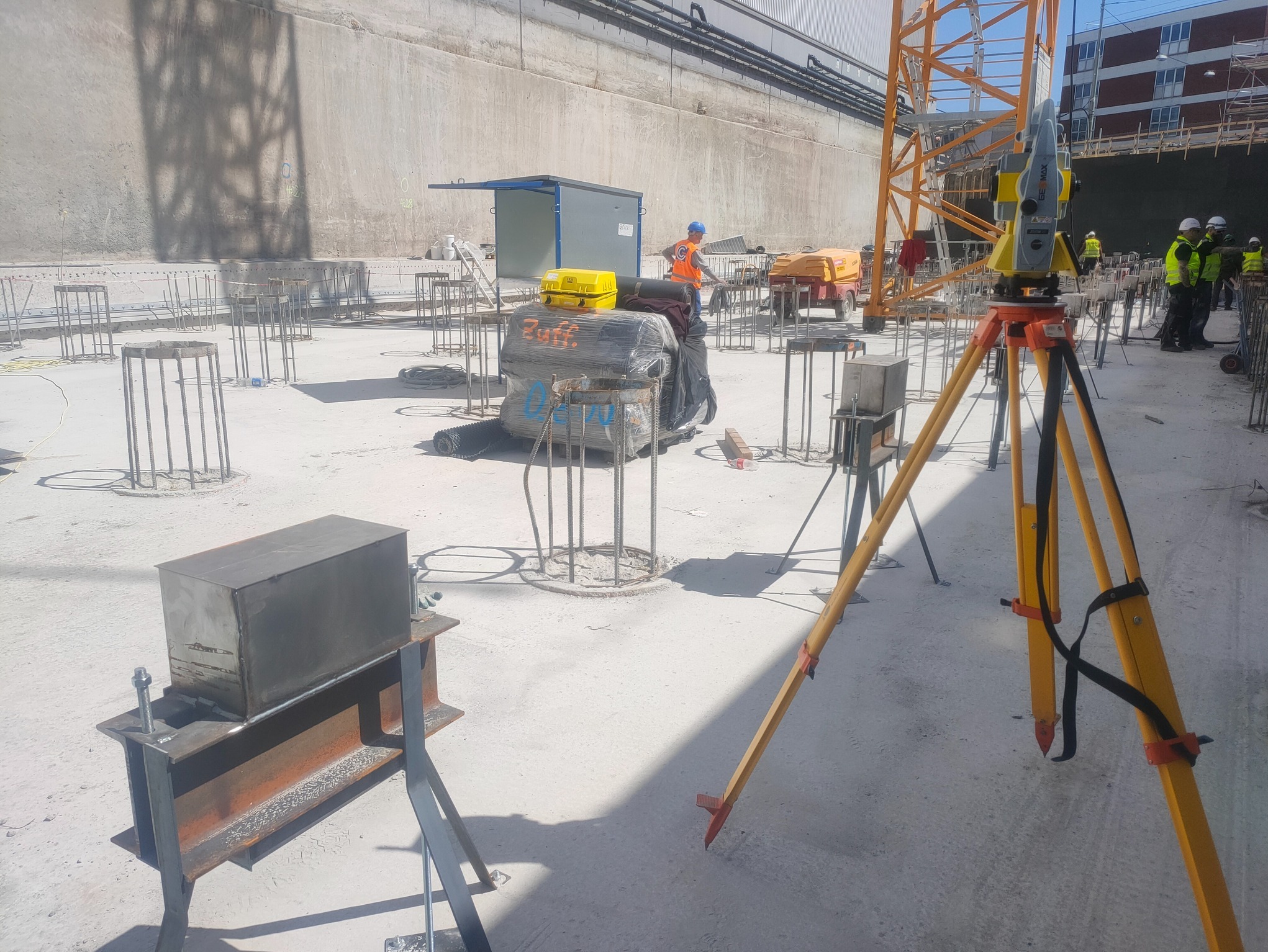 Prace geodezyjne na budowie: żółty teodolit na trójnogu, pracownicy w kamizelkach, zbrojenia i betonowa płyta fundamentowa w surowym otoczeniu placu budowy.