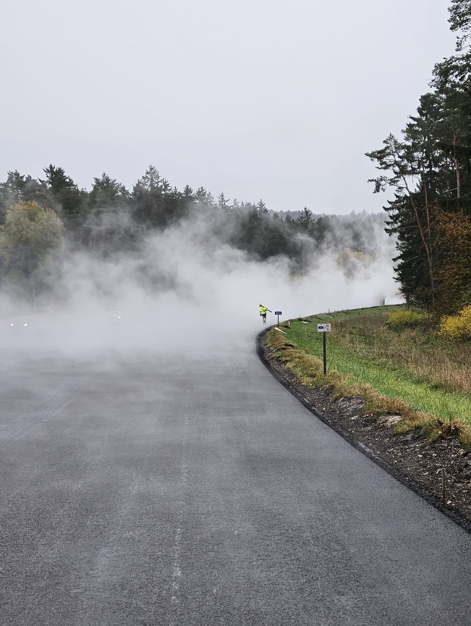 Praca na drodze w mglisty dzień: osoba w odblaskowej kamizelce z urządzeniem pomiarowym, znaki drogowe, świeżo położony asfalt i drzewa w tle.