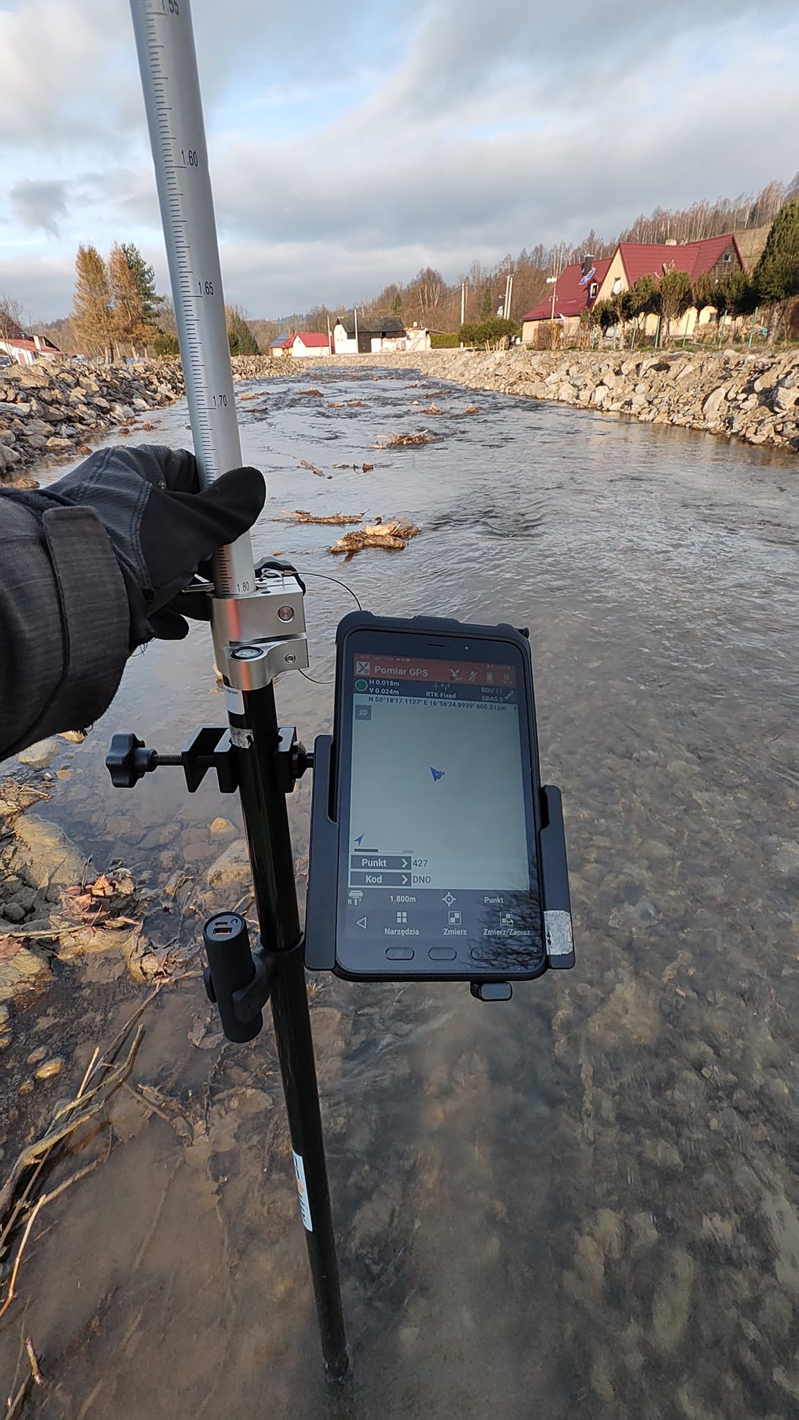 Pomiar GPS w korycie rzeki z użyciem łaty geodezyjnej i tabletu z aplikacją pomiarową. W tle zabudowa mieszkalna i drzewa.