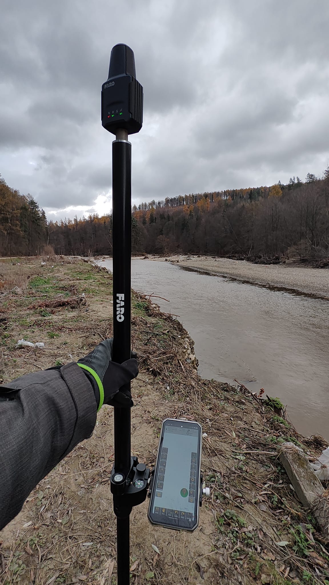 Geodeta z urządzeniem FARO i smartfonem nad brzegiem rzeki, wykonujący pomiary. Pochmurne niebo w tle. Widoczne drzewa i brzeg rzeki.
