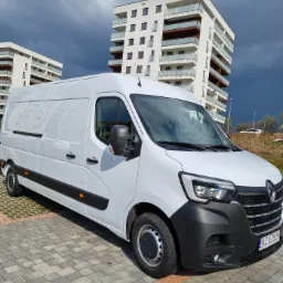 Biały samochód dostawczy Renault Master zaparkowany na kostce brukowej przed nowoczesnym budynkiem mieszkalnym w pochmurny dzień.