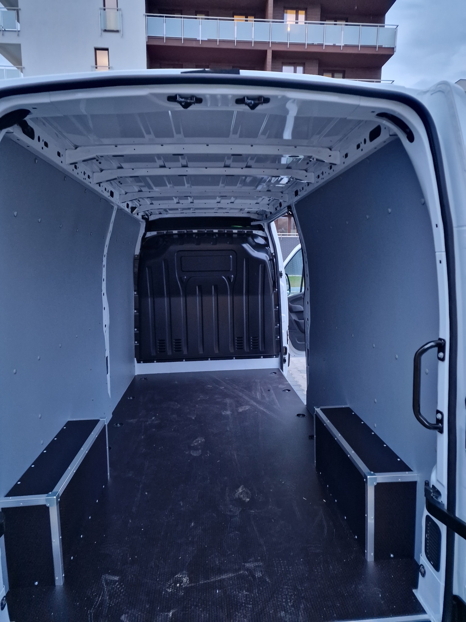 Wnętrze pustego, zabezpieczonego busa z zabudową ścian i podłogi, przygotowanego do przewozu rzeczy, widok z tyłu na tle budynku mieszkalnego.