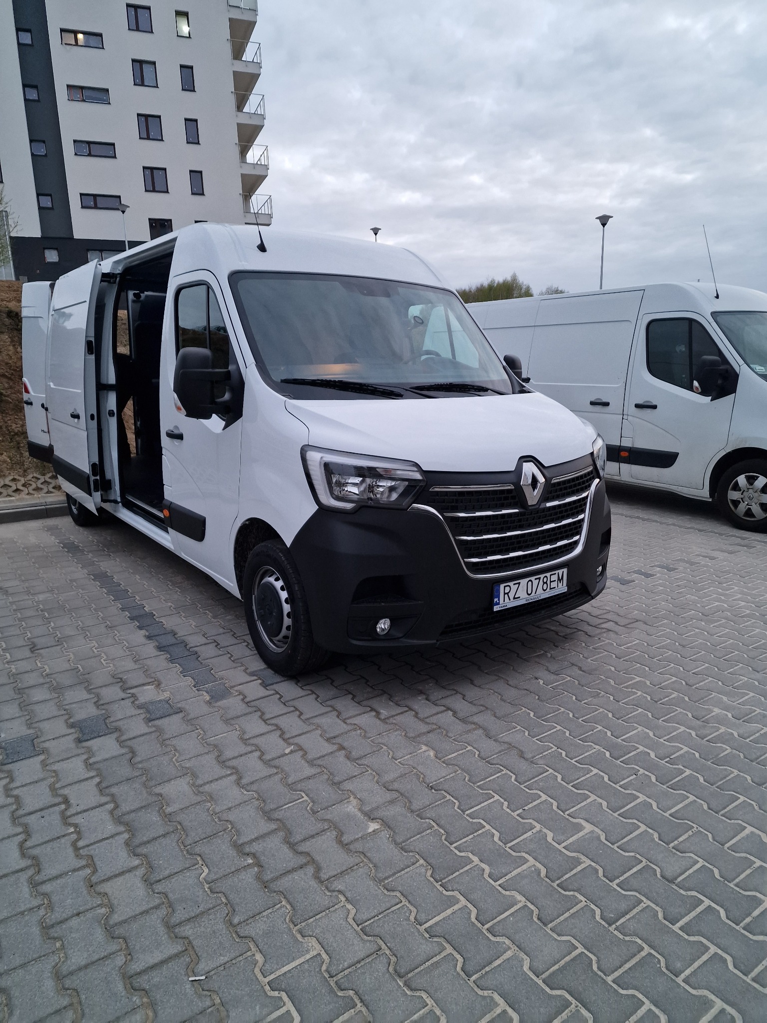 Biały bus Renault Master z otwartymi drzwiami przesuwnymi, zaparkowany na kostce brukowej przed nowoczesnym budynkiem mieszkalnym. Widoczny numer rejestracyjny RZ 078EM. W tle zaparkowany drugi...
