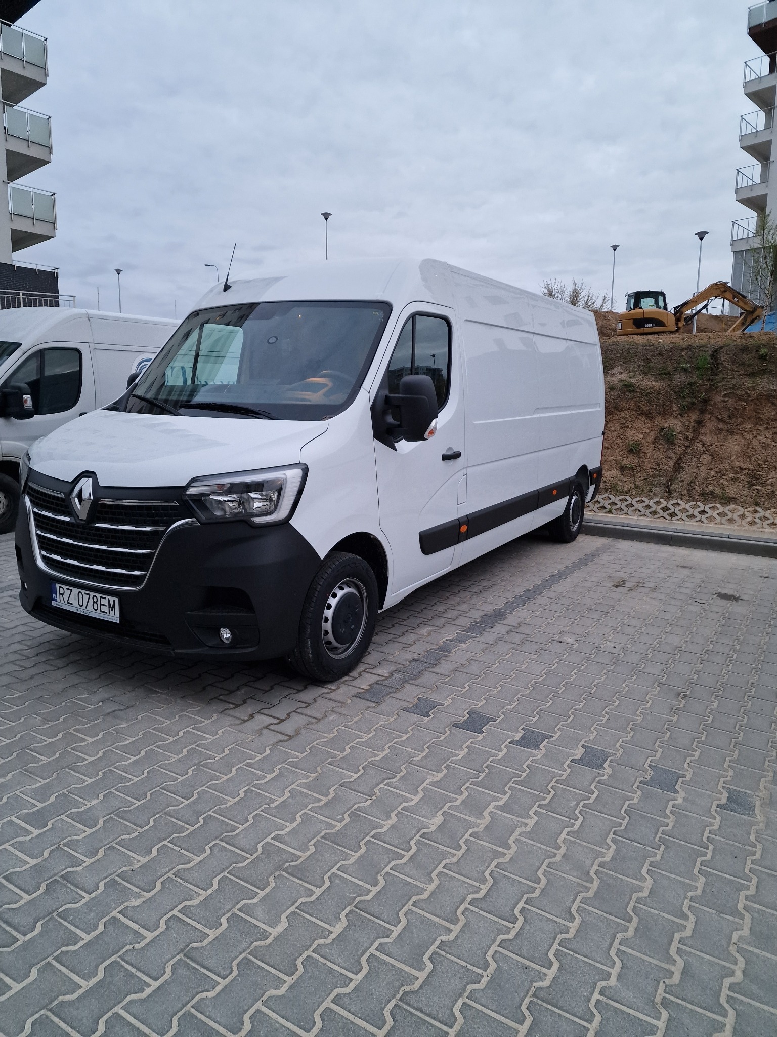 Biały bus Renault Master na parkingu z kostki brukowej, w tle budynek mieszkalny i koparka na nasypie ziemnym.