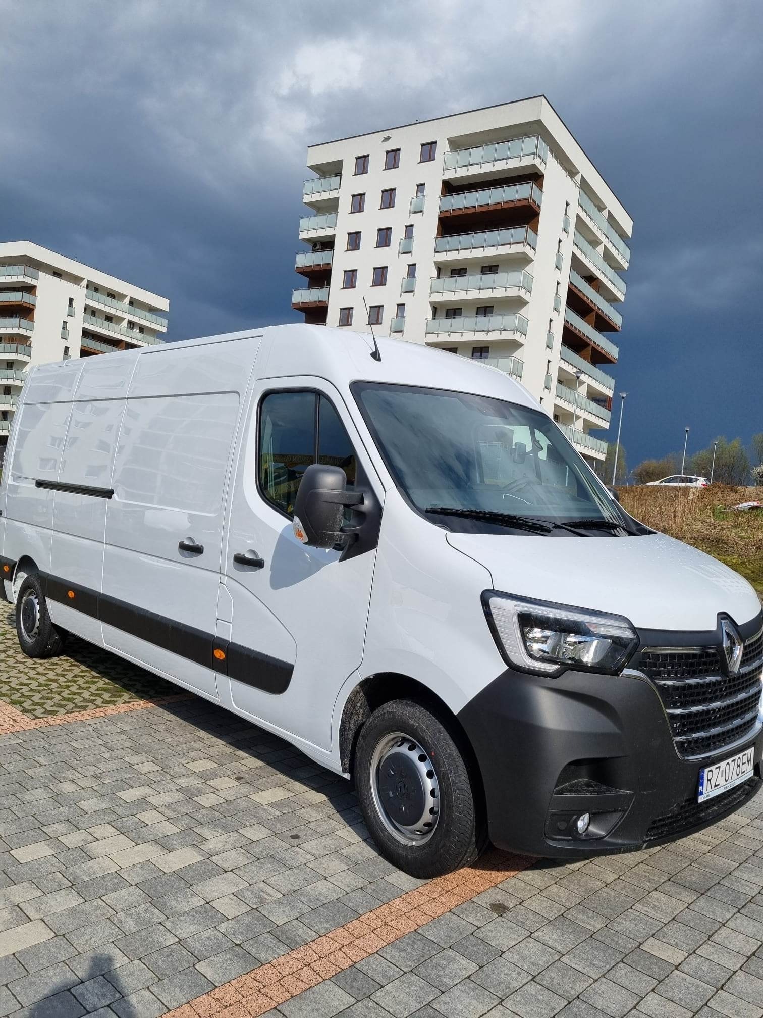 Biały samochód dostawczy Renault Master zaparkowany na kostce brukowej przed nowoczesnym budynkiem mieszkalnym w pochmurny dzień.