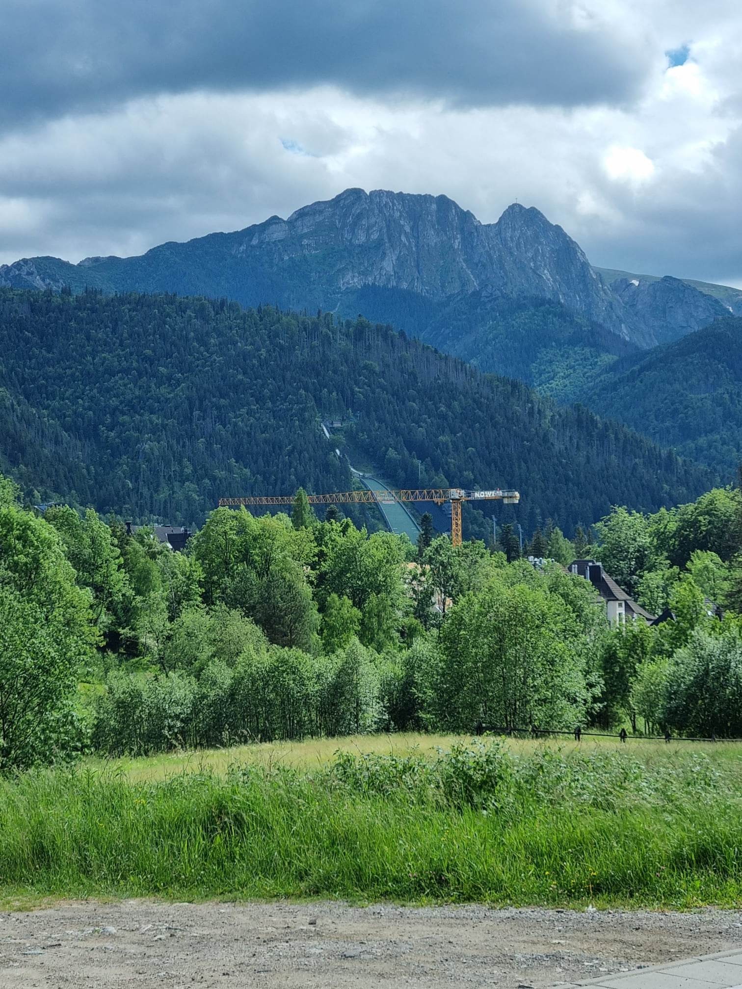 Krajobraz górski z widoczną skocznią narciarską i żółtym dźwigiem budowlanym w oddali, otoczony zielenią drzew i łąk.
