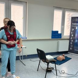 MEDYCZNE CENTRUM SZKOLENIOWE - Instruktorka demonstruje na kursantce w niebieskim stroju medycznym, jak prawidłowo wykonać manewr Heimlicha, używając specjalnego trenażera z czerwonym pasem.