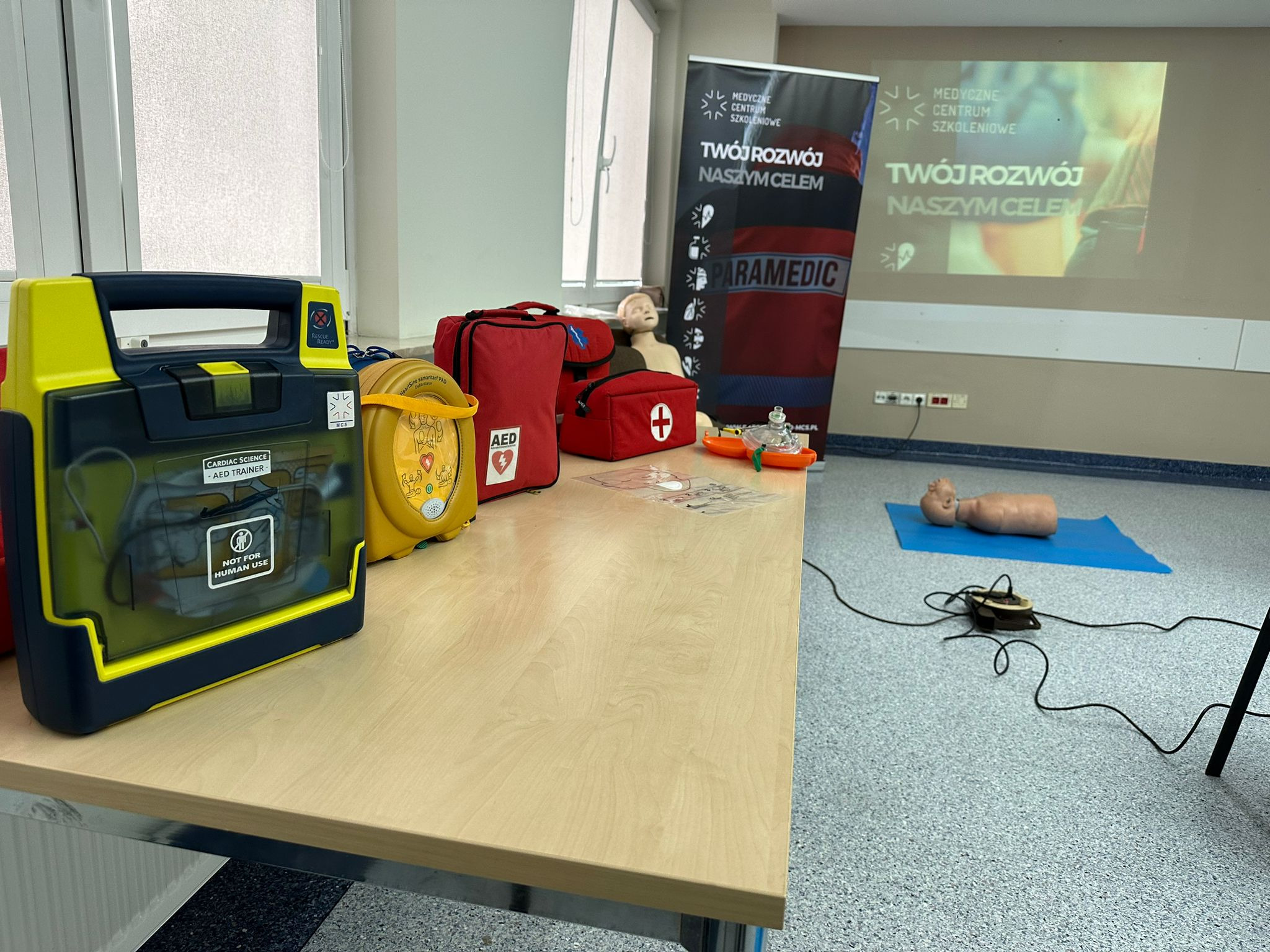 Sala szkoleniowa z fantomem niemowlęcia na niebieskiej macie, manekinem do ćwiczeń RKO na stole, automatycznymi defibrylatorami treningowymi AED i torbami medycznymi, w tle baner z logo centrum...