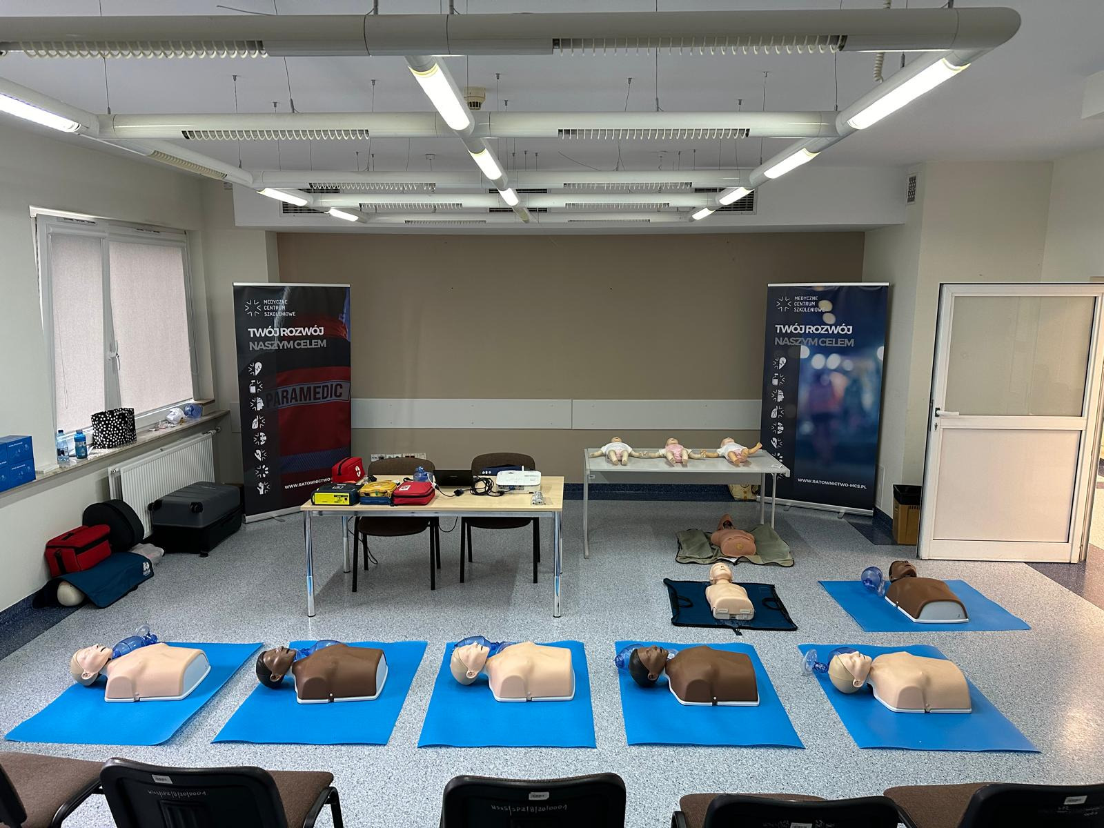 Sala szkoleniowa z manekinami do resuscytacji na niebieskich matach, fantomy niemowląt na stole, defibrylator, projektor, oraz banery reklamowe centrum szkoleniowego.