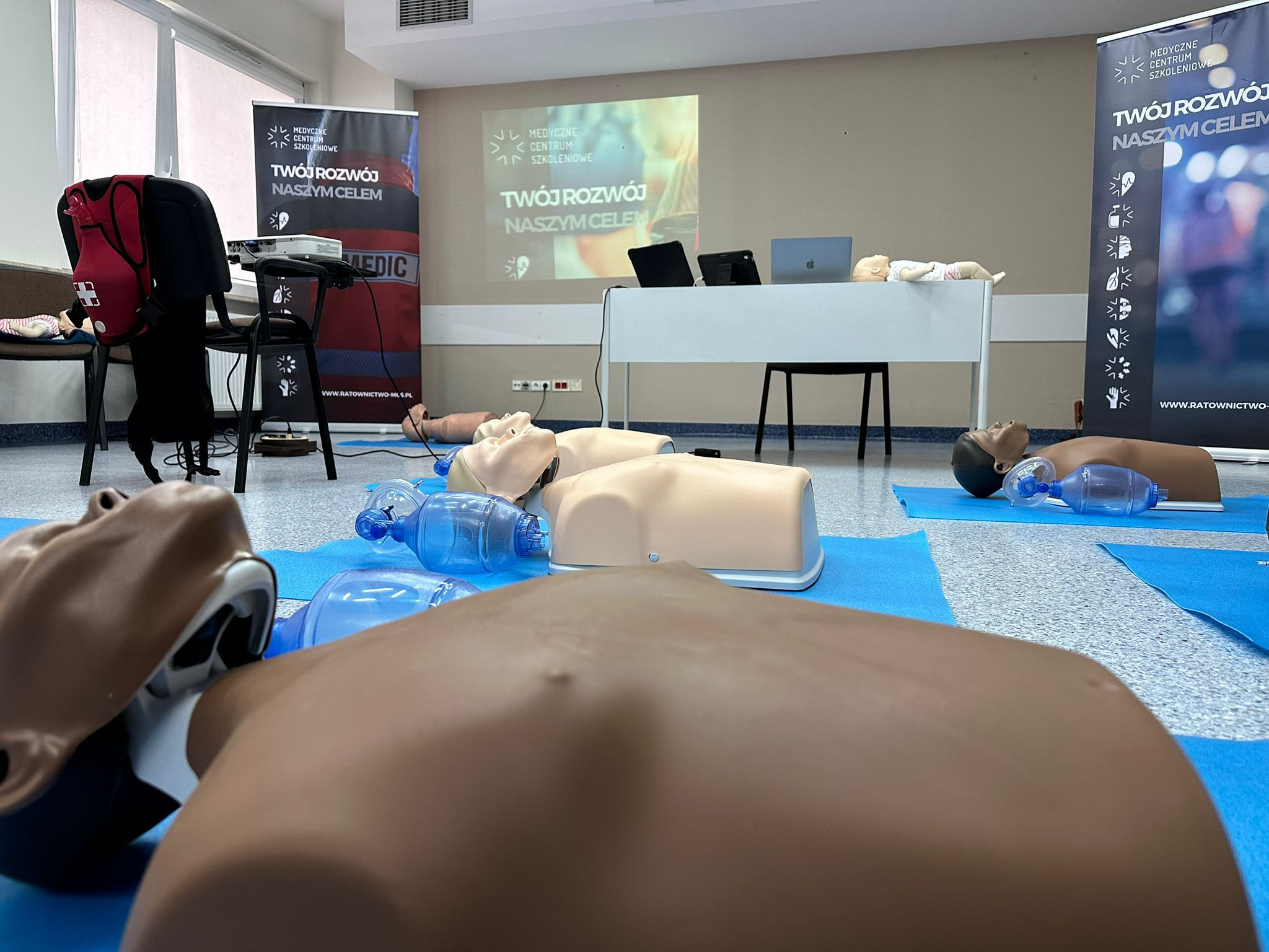 Sala szkoleniowa z manekinami do resuscytacji na niebieskich matach, widok z perspektywy jednego z manekinów, w tle rzutnik wyświetlający logo centrum szkoleniowego.