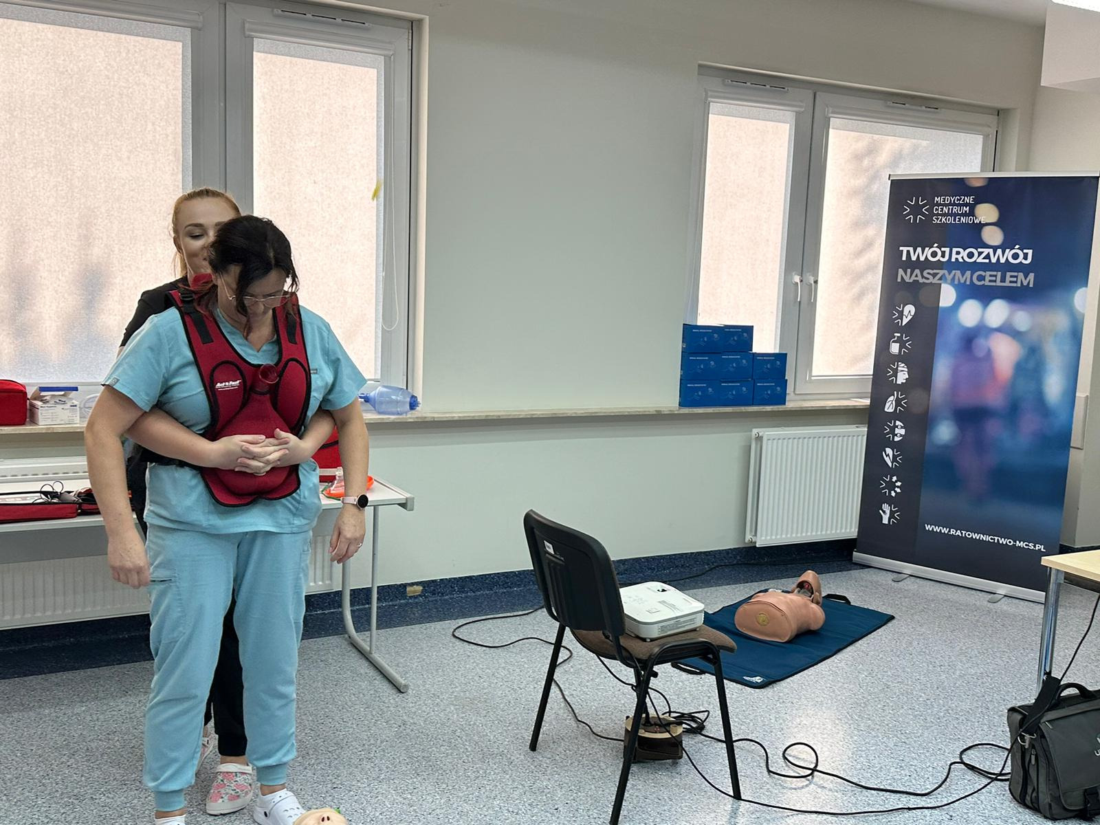 Instruktorka demonstruje na kursantce w niebieskim stroju medycznym, jak prawidłowo wykonać manewr Heimlicha, używając specjalnego trenażera z czerwonym pasem.