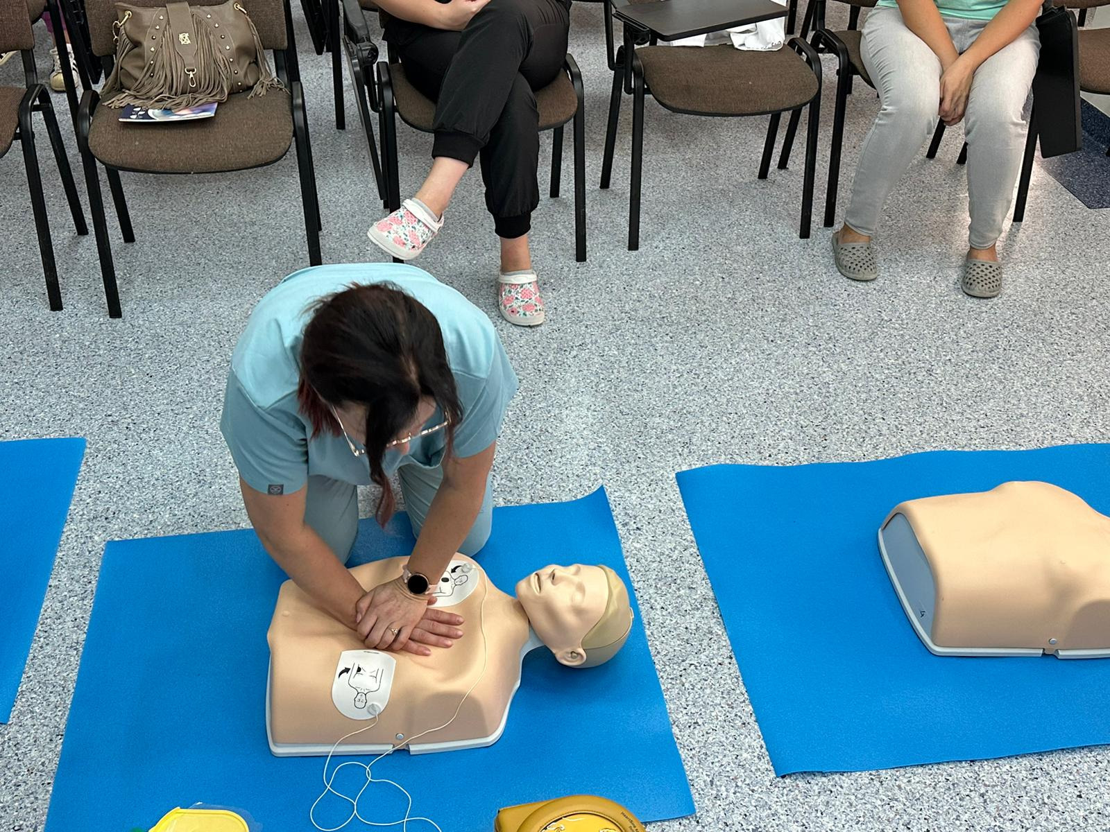 Instruktorka w jasnoniebieskim uniformie demonstruje resuscytację krążeniowo-oddechową na manekinie na niebieskiej macie, z uczestnikami kursu w tle siedzącymi na krzesłach.