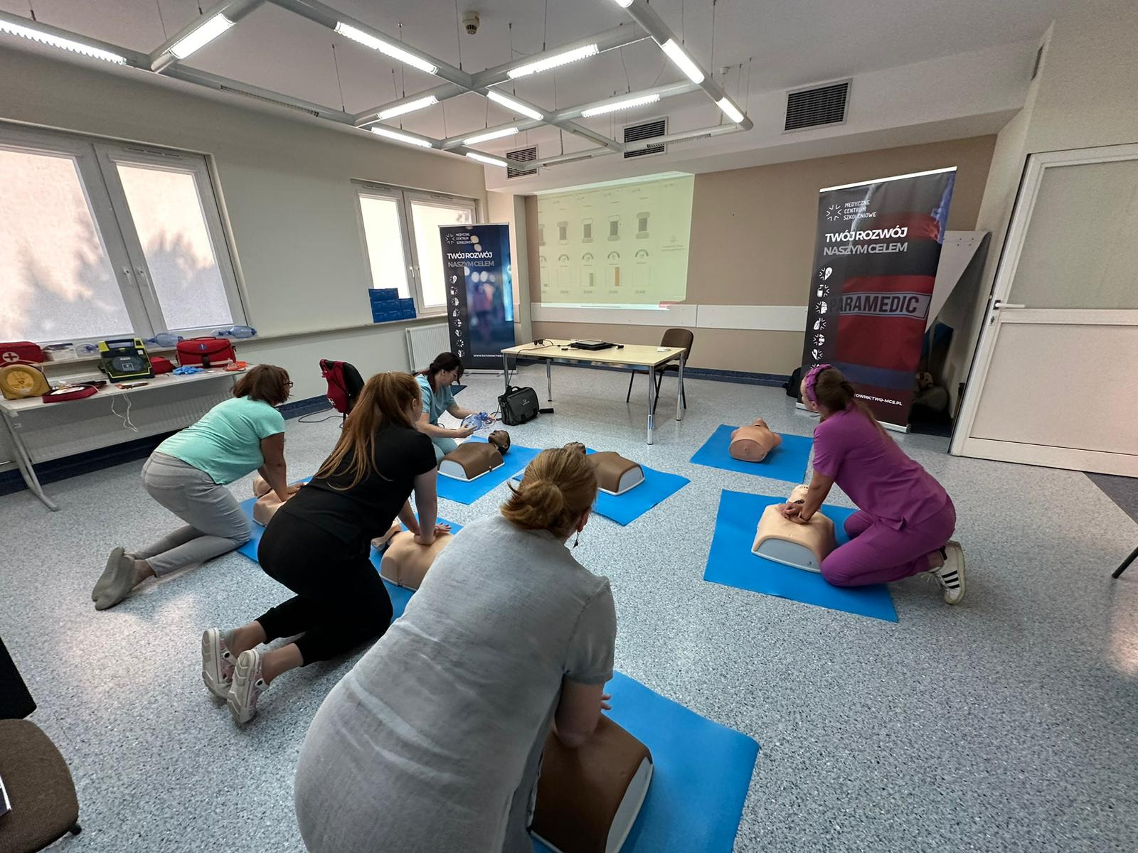 Grupa osób ćwiczy resuscytację krążeniowo-oddechową na fantomach w sali szkoleniowej, rzut ukośny z widocznym projektorem i bannerem reklamowym.